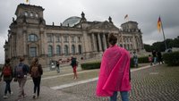 A woman with a pink shawl around her shoulders is standing in front of the German parliament building. 