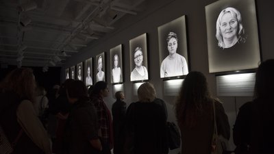 n einer dunklen Ausstellungshalle sind beleuchtete Schwarz-Weiß-Porträts von Frauen zu sehen.