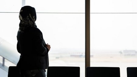Eine Frau steht im Wartebereich eines Flughafens und blickt auf das Rollfeld.