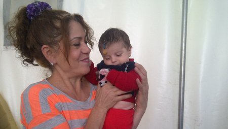 Eine Frau in einem Zelt in einem Geflüchtetenlager hält ein Baby auf dem Arm und lächelt es an. 