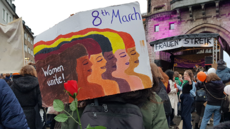 Zahlreiche Menschen beim Frauenstreik in Köln, im Vordergrund ein Demo-Plakat mit verschiedenen gemalten Frauengesichtern im Profil und dem Aufruf Women Unite