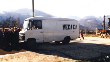A van converted for use as a medical clinic is parked at the side of a road in a rural area of Bosnia. A large number of women are waiting in front of it.