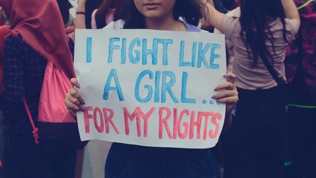 A woman is holding a placard with a hand-painted slogan “I fight like a girl for my rights” towards the camera, only the lower part of her face below her nose is visible. In the background, more women are demonstrating. 