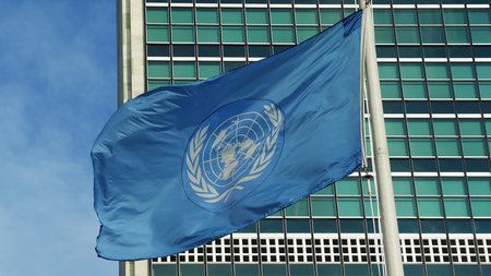 Die UN-Flagge weht an einem Masten, im Hintergrund die Fassade eines Hochhauses. 