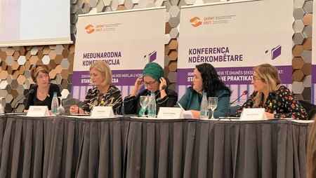 Fünf Frauen sitzen an einem langen Tisch. Im Hintergrund sind Plakate mit dem Titel der Konferenz zu sehen.