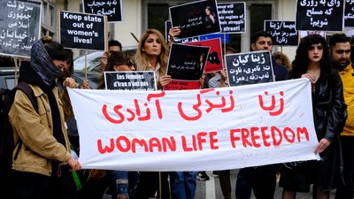 Women are holding a white banner with the words “Woman. Life. Freedom” in Persian and English painted in red on it.