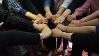 A group of women are standing in a circle, they have each clenched a fist and are holding these together in a small circle in their centre.