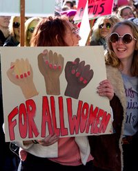 Several women at a Women's March hold a banner with the words For all women, above which are painted three clenched fists in different skin colours and with red fingernails.