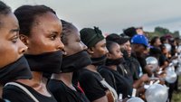 A long row of women at a demonstration in Liberia. They are standing side-by-side, their mouths are covered with a black cloth and they are holding candles.