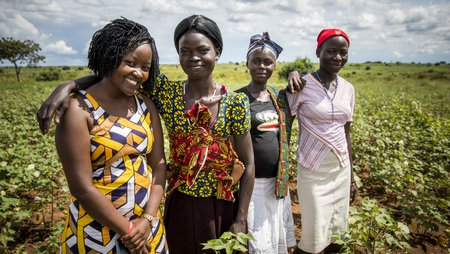 Four women are standing arm-in-arm, smiling into the camera, with green Ugandan countryside in the background.