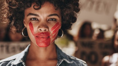 : A photo of a female activist with face paint depicting a red handprint covering her mouth and lower half of her face.