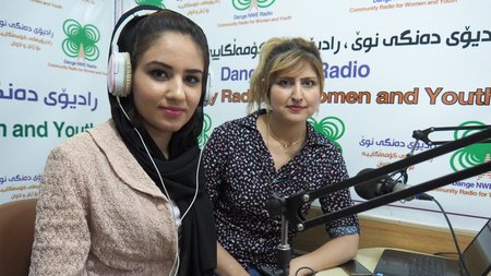 Zwei Frauen befinden sich in einem Tonstudio eines Radiosenders und sitzen vor einer Art Mischpult. Eine hat Kopfhörer auf und beide haben jeweils ein Mikrofon vor sich. 