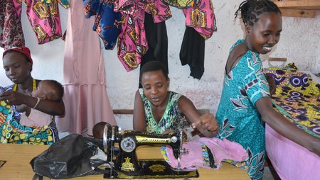 Three women participating in a sewing course. In the foreground is a mechanical sewing machine and in the background are colourful textiles.