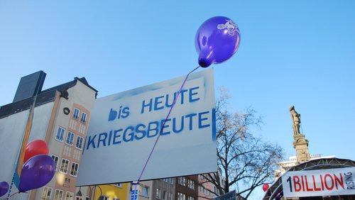 Demonstrationsplakat mit der Aufschrift “Bis heute Kriegsbeute”, im Hintergrund ein Banner von One Billion Rising.