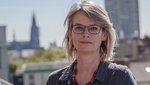 A woman wearing a dark jacket and glasses is standing on a roof terrace with Cologne Cathedral in the background. Portrait photo by Kirsten Wienberg, Head of Evaluation and Quality Department 