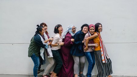 Seven women laughing and joking as they stand in a line, one in front of the other, alongside a light-grey concrete wall. They are posing for a group photo and appear to be having fun. 