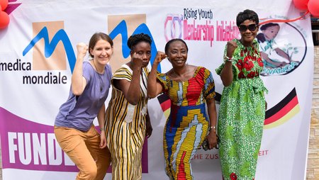 Four women with raised fists look cheerfully and confidently into the camera, in the background a poster with the logos of various women's rights organisations. 