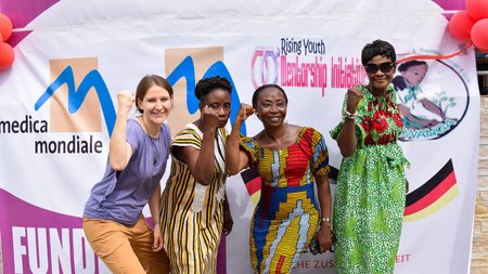 Four women with raised fists look happily and confidently into the camera, in the background a banner with the logos of various women's rights organisations.