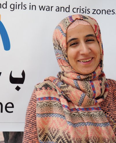 The logo of the women's rights organisation medica mondiale can be seen in the background with Arabic characters underneath. On the right in front of it is the face of a friendly smiling woman. It is legal advisor Jihan Abas Mohammed.