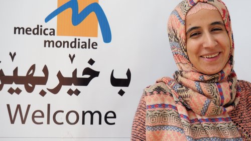 The logo of the women's rights organisation medica mondiale can be seen in the background with Arabic characters underneath. On the right in front of it is the face of a friendly smiling woman. It is legal advisor Jihan Abas Mohammed.