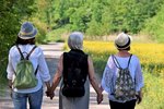 Drei ältere Frauen sind von hinten zu sehen, sie spazieren in der Natur und halten sich an den Händen. 