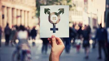 A hand is holding up a piece of paper which bears a non-binary gender symbol that stands for diversity, in the middle of which there is an equality sign. A busy pedestrian area can be seen in the background. 