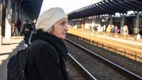 Eine ältere Frau mit einer Ukraine-Flagge an ihrem Rucksack steht an einem Bahnhof.