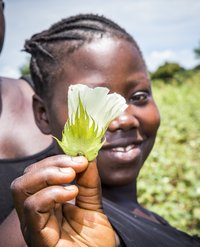 Ein Mädchen auf einem Baumwollfeld hält eine Blüte in die Kamera und lächelt.
