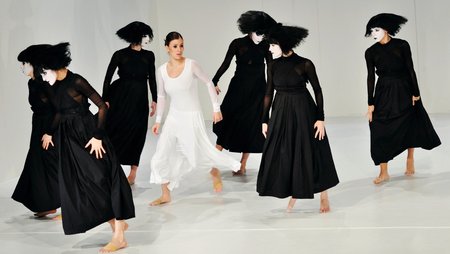 Theaterszene mit einer in weiß gekleideten Frau und mehreren in schwarz gekleideten Personen die sie bedrohlich umkreisen.
