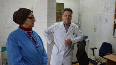 A woman and a man in white coats are standing in a hospital room talking