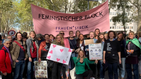 Gruppenfoto von zahlreichen medica mondiale Mitarbeiterinnen auf einer Demo, im Hintergrund ein großes Plakat mit der Aufschrift Feministischer Block Deauen*Streik