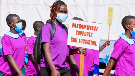 Several people are walking down a street with placards. One of them is holding a sign saying Memprow supports girl education. 