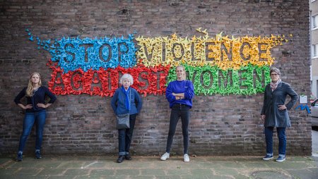 A wall bearing lettering made out of origami birds forming the words “Stop violence against women”. Four women are standing in front of the wall in poses demonstrating self-confidence.