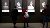 Inside an exhibition hall, visitors are looking at four large-format black-and-white photos with explanatory text beneath them. Each is a portrait photo of one woman or girl. 