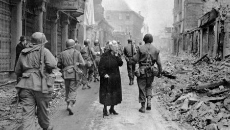 Schwarz-weiß Foto 1945: Eine alte Frau, in schwarz gekleidet, steht in der Mitte einer Straße, sie blickt gedankenverloren ins Leere. An ihr vorbei laufen zahlreiche Soldaten. Die Gebäude entlang der Straße liegen in Trümmern.
