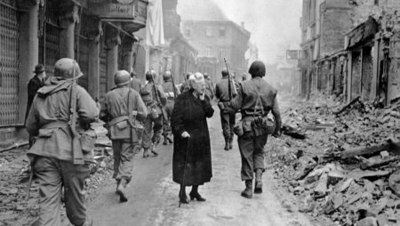 An old woman dressed in black stands in the middle of a street, lost in thought, gazing into space. Numerous soldiers walk past her. The buildings along the street are in ruins.
