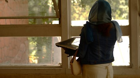 A women seen from behind. She is standing in front of a window, looking outside. She is holding a laptop in her arms..