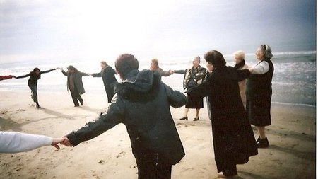 Eine Gruppe von Frauen stehen am Strand in einem großen Kreis und halten sich an den Händen, im Hintergrund das Meer und die Sonne. 