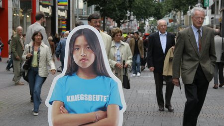 A cardboard cut-out of a girl wearing a T-shirt with the slogan “Spoils of War”. This figure is standing in the pedestrian zone in Cologne city, with people walking quickly by.