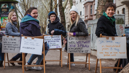 medica mondiale Mitarbeiterinnen sitzen auf Stühlen in einer Kölner Straße, an den Stühlen hängen Plakate mit feministischen Botschaften.