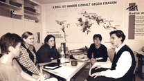 Eine Gruppe von Frauen sitzt in einer Arbeitsrunde an einem Tisch mit Schreibutensilien. Im Hintergrund ist eine Weltkarte zu sehen. 