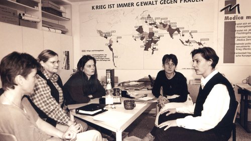 Eine Gruppe von Frauen sitzt in einer Arbeitsrunde an einem Tisch mit Schreibutensilien. Im Hintergrund ist eine Weltkarte zu sehen. 