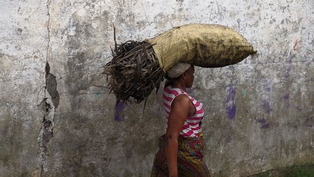 A woman is carrying a sack of brushwood on her head as she walks past a grey, cracked wall.
