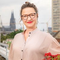 Eine Frau in einer hellen Bluse und mit rotem Lippenstift steht auf einer Dachterrasse mit Kölner Dom im Hintergrund. Portraitfoto von Kristina Remy, Leitung Finanzen und Verwaltung bei medica mondiale.
