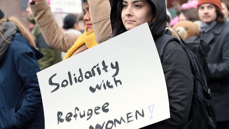 A large number of people at a demonstration, in the foreground a woman is standing with a poster bearing the words “Solidarity with refugee women!”