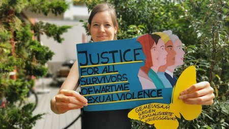A woman is holding a hand-drawn poster with the slogan “Justice for all survivors of wartime sexual violence. “Gemeinsam gegen sexualisierte Kriegsgewalt.”