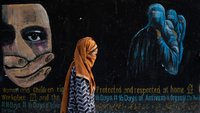 A woman wearing a brown headscarf passes a darkly painted wall showing protest phrases