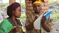 Zwei Mitarbeiterinnen der kongolesischen Frauenrechtsorganisation PAIF diskutieren über den Inhalt eines Dokuments, das eine der beiden in Händen hält. Im Hintergrund ist eine ländliche Gegend zu sehen.  