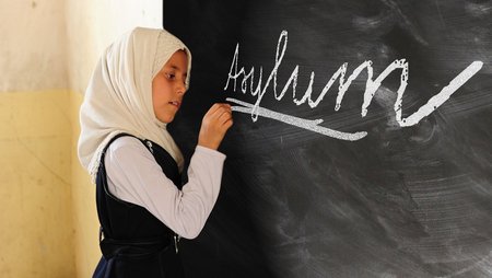 Ein junges Mädchen mit Kopftuch schreibt mit Kreide das war Asylum an eine Tafel. 