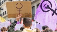 A man seen from behind holds a placard with the venus sign. He is joining a demonstration. Right in the picture is a purple flag showing the venus sign with a raised fist.